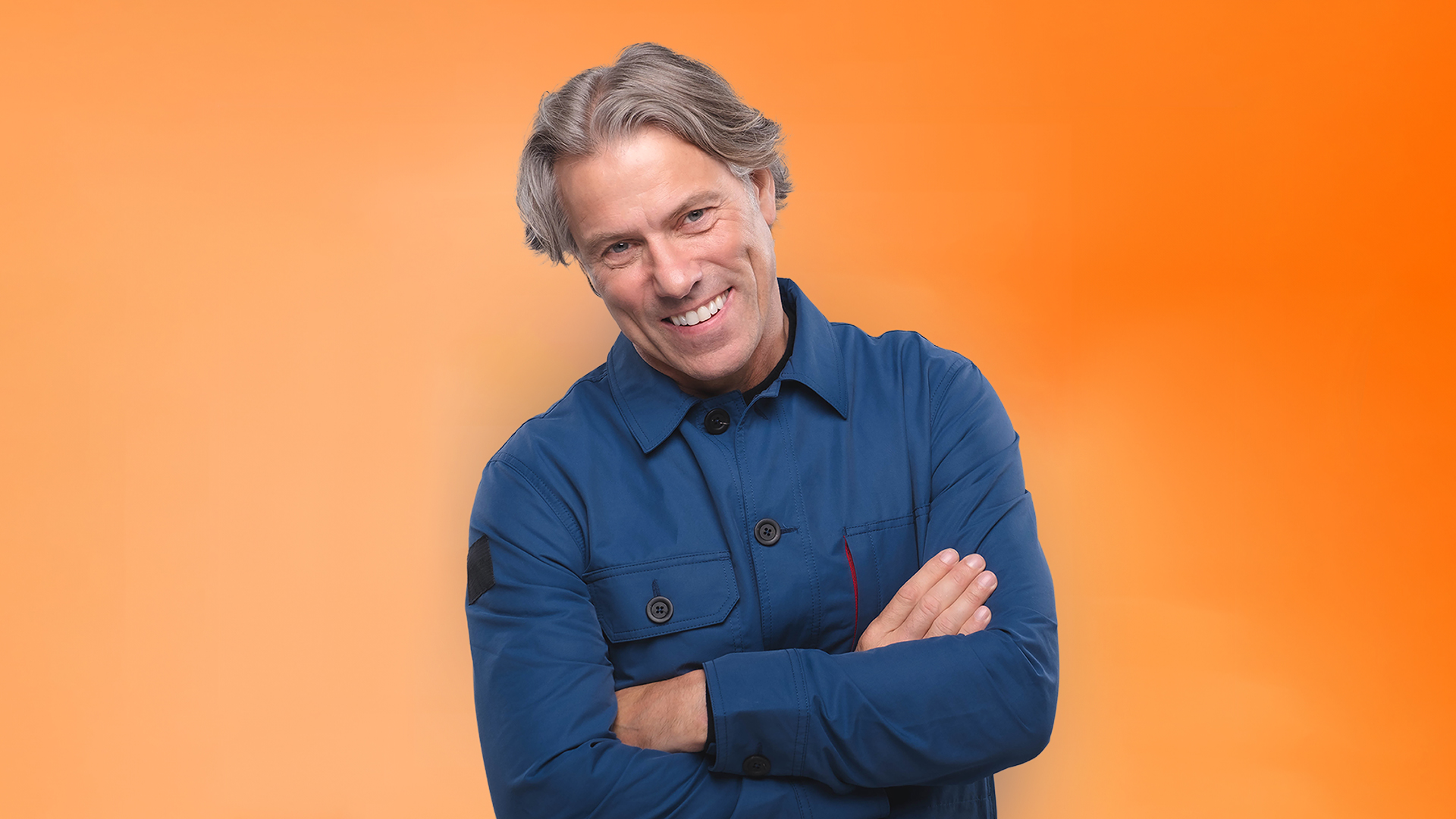John Bishop pictured from waist up. He wears a blue overall style top and stands smiling with his arms folded. Orange backdrop.