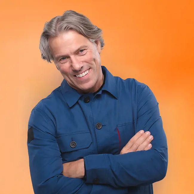 image John Bishop pictured from waist up. He wears a blue overall style top and stands smiling with his arms folded. Orange backdrop.