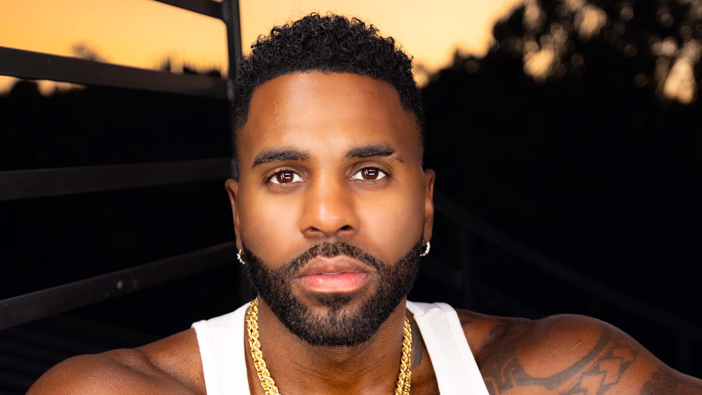 Jason Derulo looks straight to camera. Close up of Jason's head and shoulders. He wears a white vest with yellow gold jewellery and has a sunset backdrop
