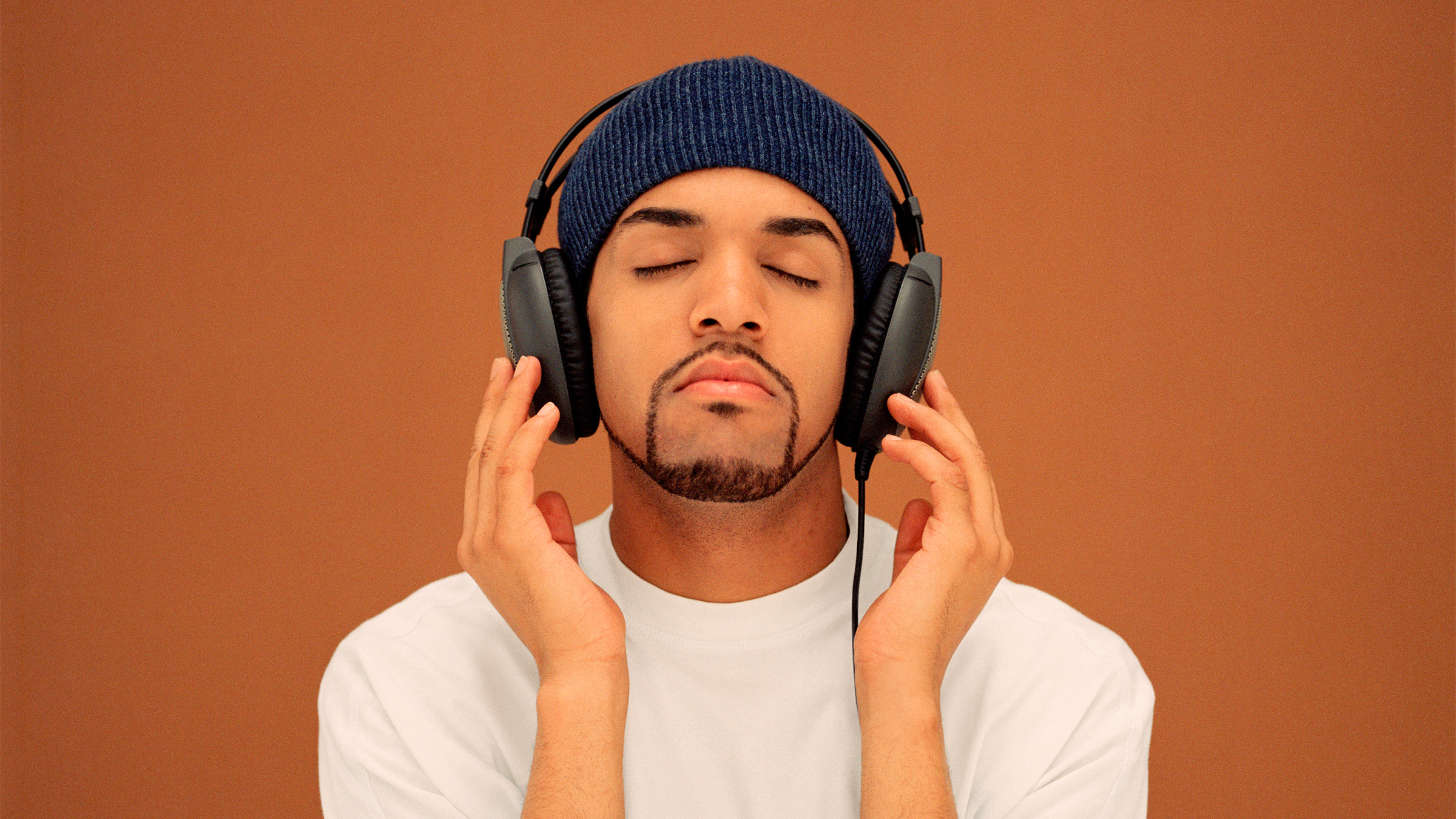 Craig David headshot wearing a dark blue beanie hat, white T-shirt and headphones. His eyes are closed seemingly listening to music