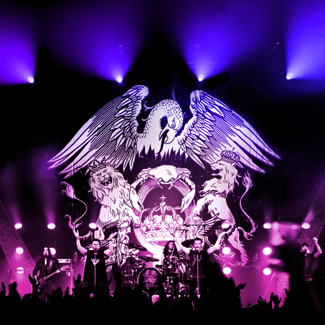 Queen Extravaganza stage image taken from a distance. A shot of the band on stage with blue lights above and purple stage lights. Crest with eagle and two lions as backdrop.