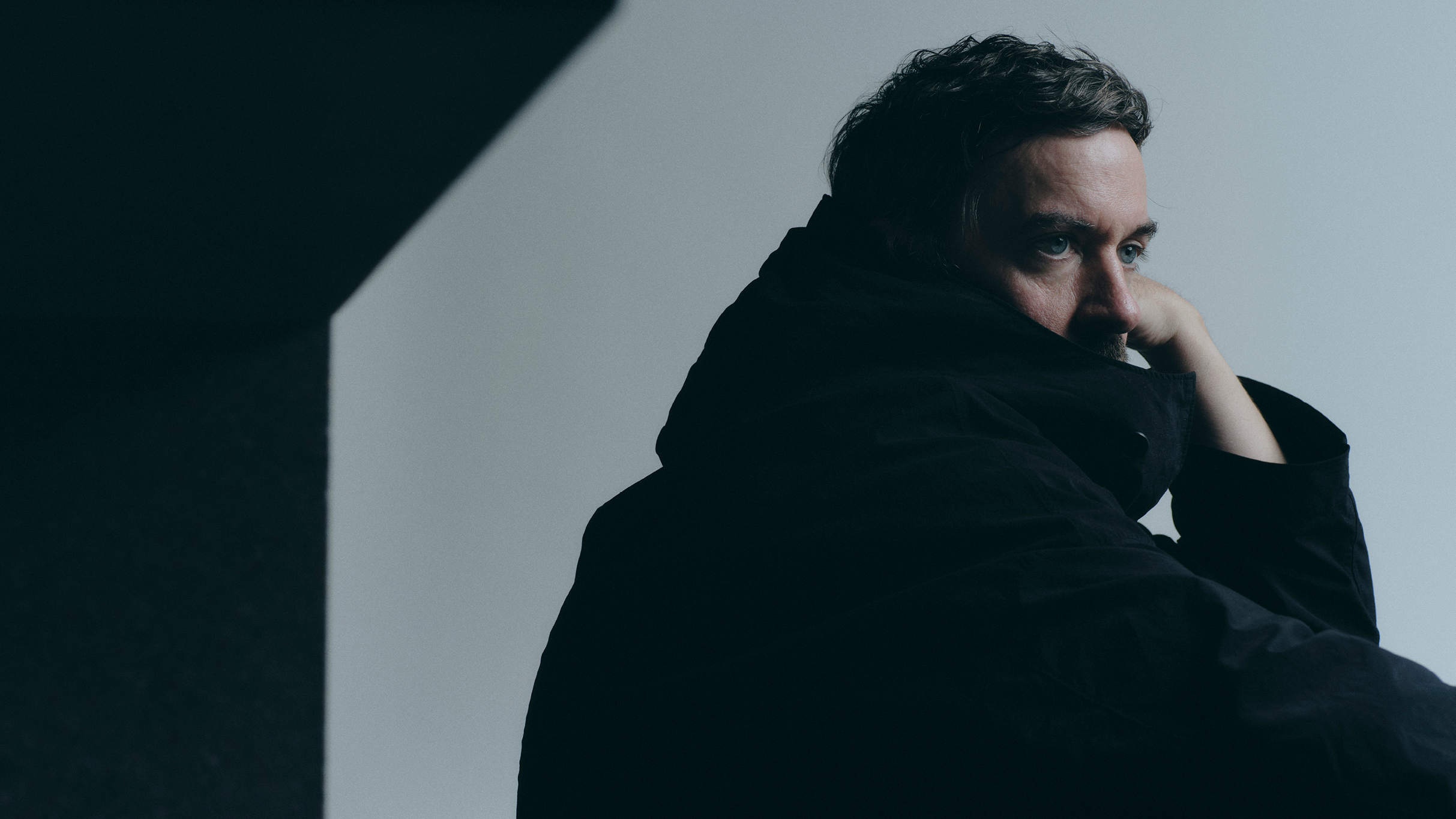 Bonobo press shot. Profile of artist sat resting head on hand. Wearing heavy, high collared black hoodie which masks lower part of his face.