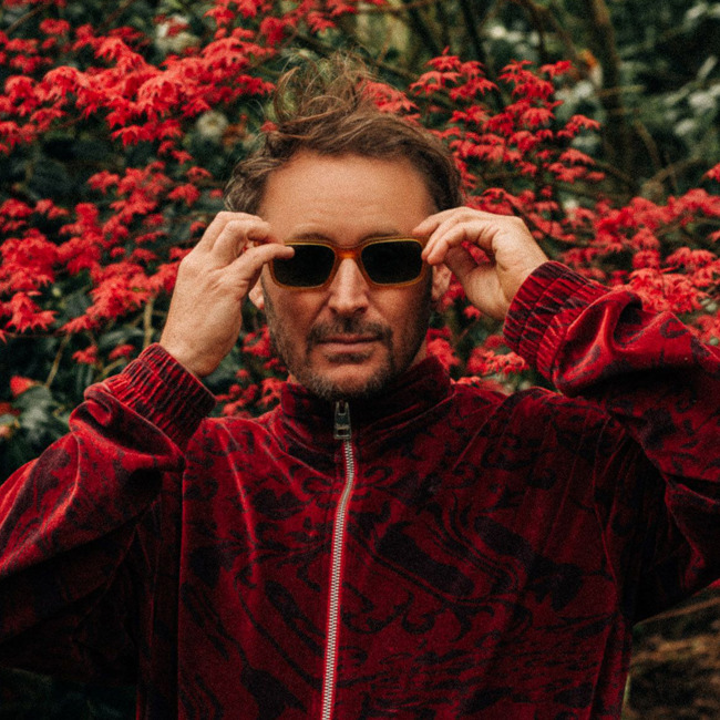 Official shot of Ben Howard pictured putting on sunglasses, looking direct to camera, wearing a red zipped velour jacket in front of a red leafed Japanese Maple tree.